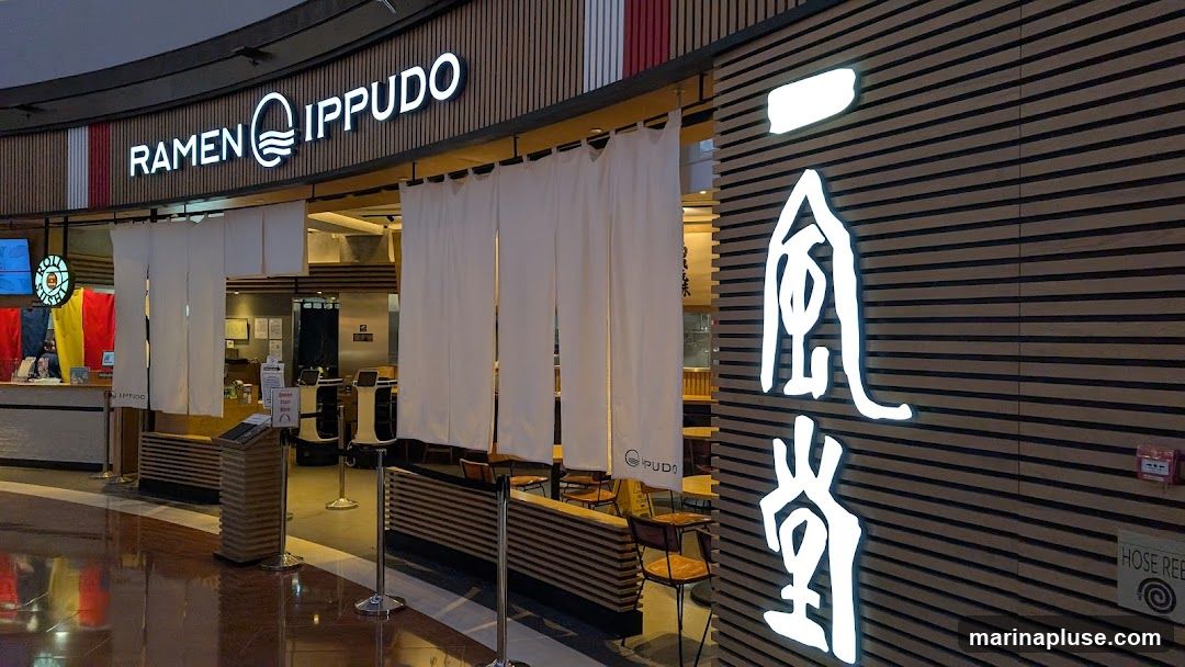 The exterior of a Ramen Ippudo restaurant. The storefront features a prominent sign with the restaurant's name and logo. White curtains hang across the entrance, and a large Japanese character is illuminated on the wooden facade. Inside, tables and chairs are visible, along with a counter and some robotic servers.