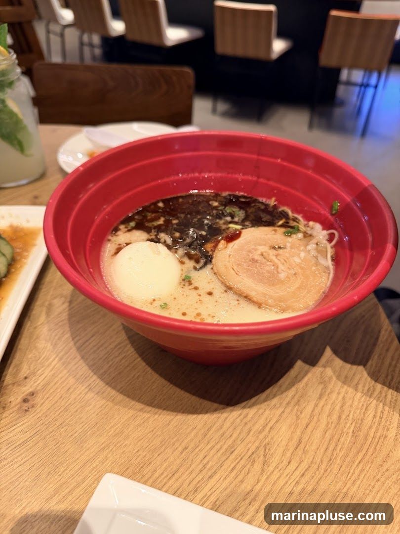 A close-up shot of a red bowl filled with ramen. The ramen includes broth, noodles, a soft-boiled egg, and a slice of chashu pork. In the background, out of focus, are chairs and tables, suggesting a restaurant setting. To the left, a glass of lemonade with mint and ice is visible.