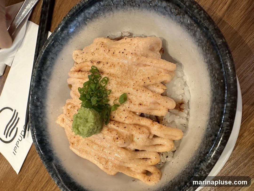 A close-up overhead shot of a salmon donburi dish. The dish features white rice topped with a generous portion of grilled salmon coated in a creamy sauce, garnished with chopped green onions and a dollop of wasabi. A napkin with the logo 'IPPUDO' is visible to the left of the bowl.