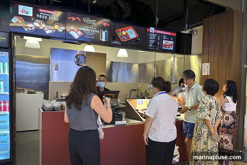 A food stall with customers ordering at the counter. Digital menus display bento boxes, a special offer for a free herbal drink with purchase, and a list of menu items. A staff member in a mask is serving customers.