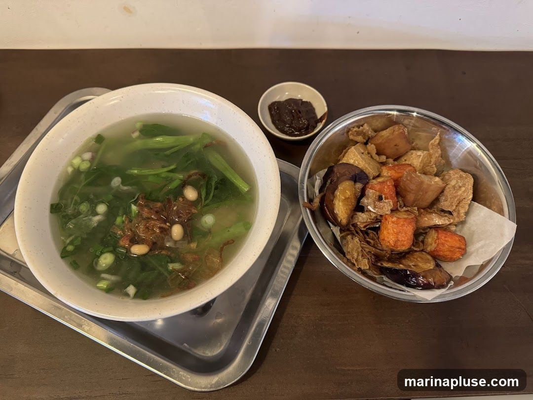 A bowl of clear vegetable soup with greens, scallions, fried shallots, and beans is served on a metal tray. Next to it is a metal bowl filled with various fried items including eggplant, tofu, and what appear to be fried dough sticks. A small dish of dark sauce is also visible.