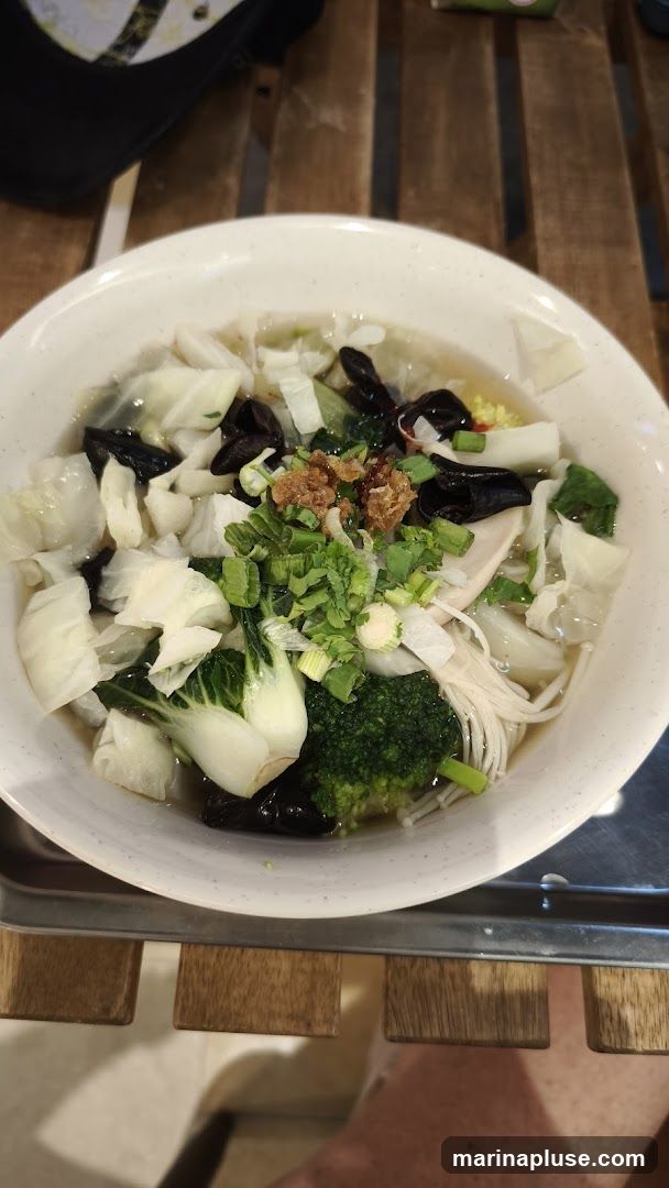 A close-up shot of a white bowl filled with a clear broth soup. The soup contains various vegetables including napa cabbage, bok choy, broccoli florets, enoki mushrooms, wood ear mushrooms, and chopped green onions. Crispy fried shallots are sprinkled on top as a garnish. The bowl is placed on a wooden slatted surface.