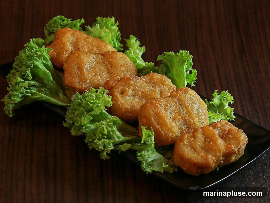 A close-up shot of chicken nuggets served on a bed of lettuce on a black plate. The nuggets are golden brown and appear crispy. The background is a dark wooden surface.