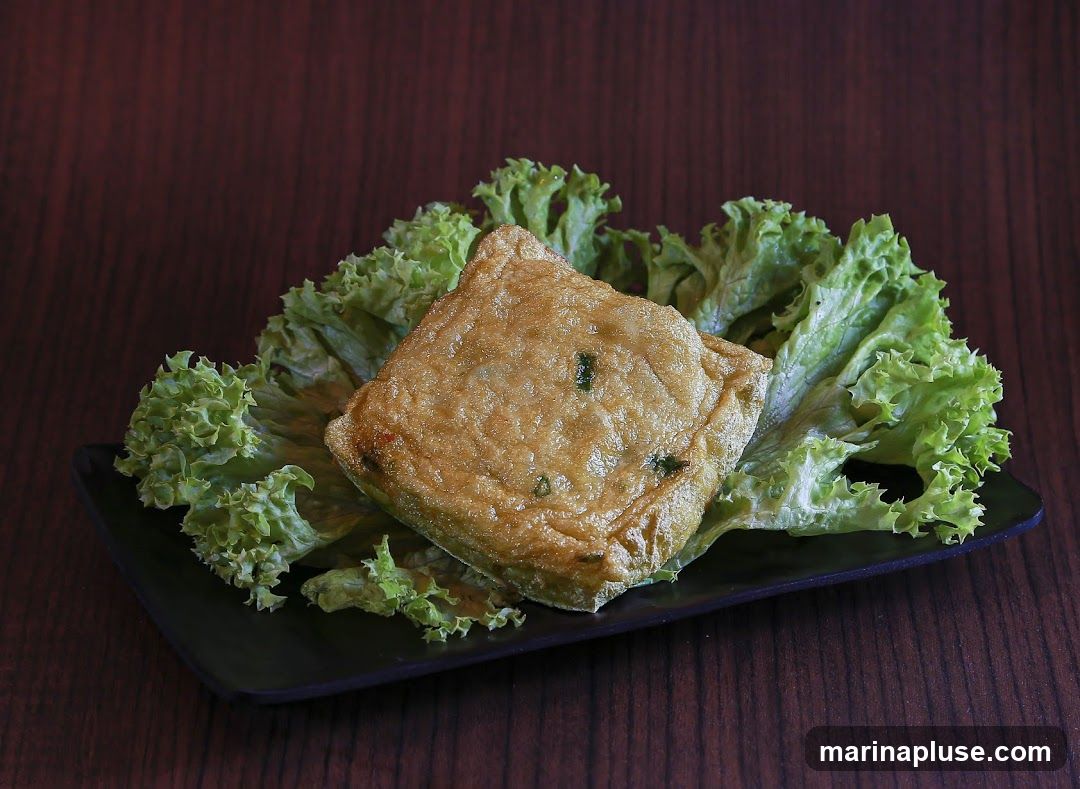 A square piece of golden-brown fried tofu, garnished with green onions, is served on a bed of fresh green lettuce leaves. The dish is presented on a black rectangular plate, set against a dark wooden background.