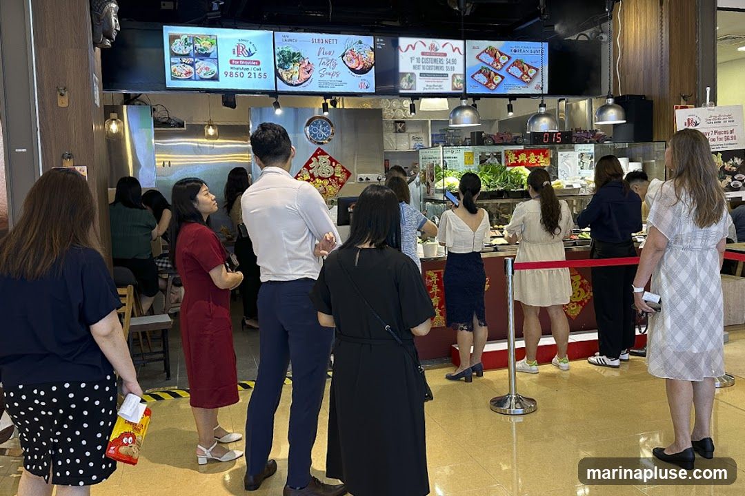 A group of people are standing in line at a food court. Digital menu boards above display various food items and promotions. One board shows a phone number for inquiries. Another advertises a new soup launch and a Korean bento box. A timer shows '015'. Red ropes guide the queue.