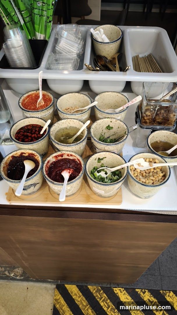A condiment bar with various small bowls filled with different toppings and sauces. There are also stacks of plastic cups, containers, spoons, chopsticks, and skewers. Some of the visible condiments include chili powder, chopped nuts, chopped herbs, chili paste, chopped scallions, and soybeans.