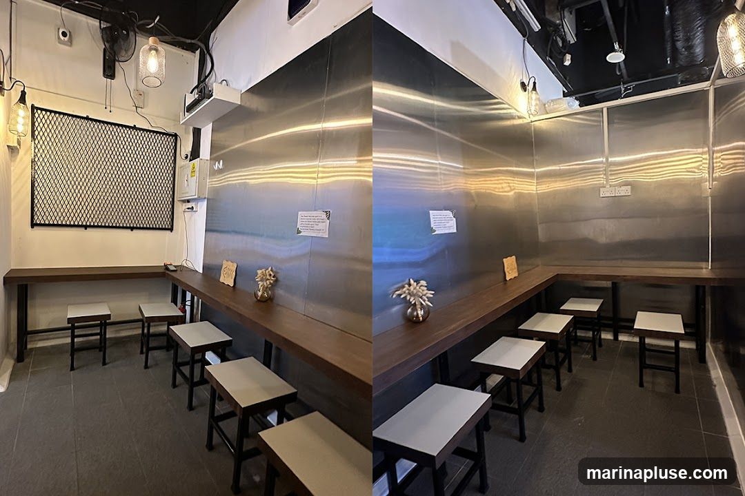 The image shows the interior of a cafe with a minimalist design. There are long wooden counters along the walls with stools for seating. The walls are covered in stainless steel panels, and industrial-style pendant lights hang from the ceiling. A black metal mesh panel is mounted on one wall. Small decorative items, such as vases with dried flowers and wooden fish cutouts, are placed on the counters. There are also signs posted on the stainless steel walls.