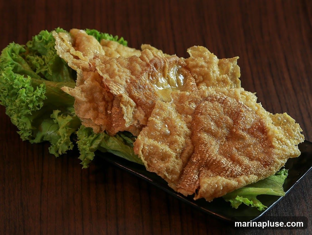 A close-up shot of a golden-brown, crispy fried dish served on a bed of fresh green lettuce. The dish appears to be thin and flaky, with a slightly oily sheen. It is presented on a dark rectangular plate against a dark wooden background.