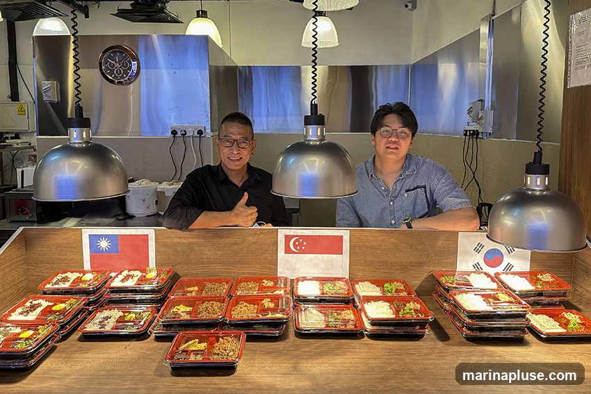 Two men are standing behind a wooden counter filled with pre-packaged bento boxes. The man on the left, wearing glasses and a black shirt, gives a thumbs-up. The man on the right, wearing glasses and a denim shirt, looks towards the camera. In front of them, flags of Taiwan, Singapore, and South Korea are displayed. The bento boxes appear to contain rice, meat, and vegetables. The background shows a stainless steel kitchen area with hanging lamps and a clock.