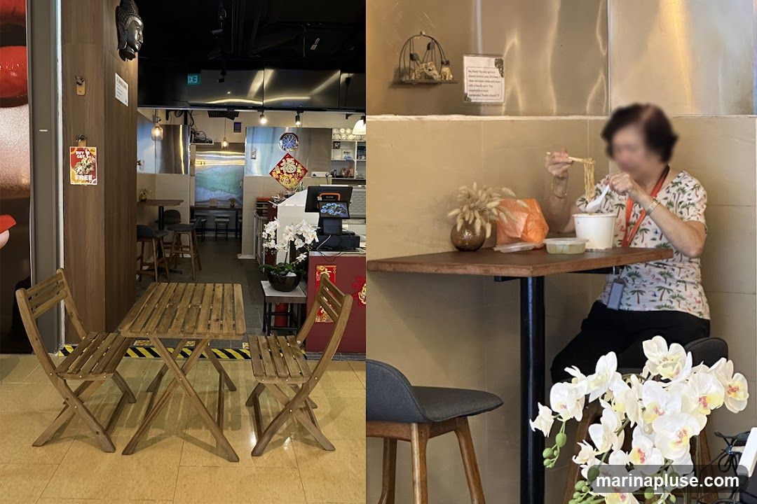 The image shows the interior of a restaurant. On the left, there is an empty wooden table and chairs set up. The background reveals a counter area with kitchen equipment and decorations. On the right, an elderly woman is seated at a table, eating noodles from a bowl. White orchids are visible in the foreground.
