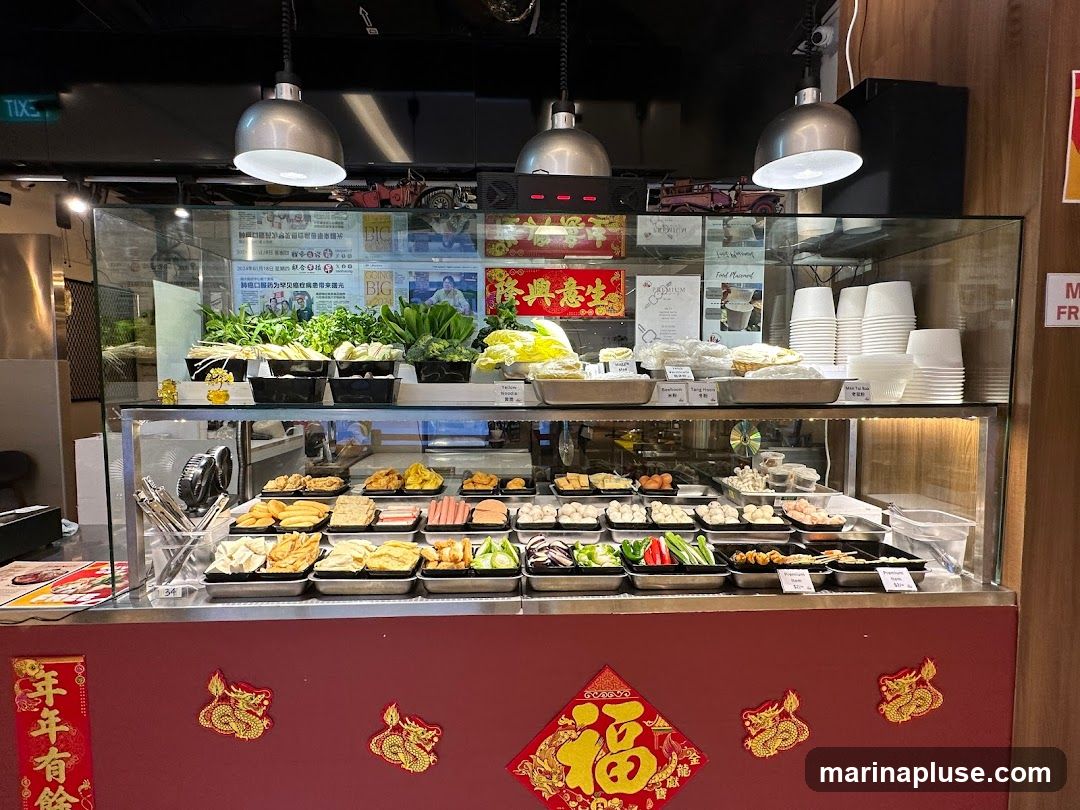 A refrigerated display case filled with various ingredients for hot pot or a similar dish. The top shelf holds fresh vegetables like leafy greens, broccoli, and herbs. Below, numerous trays contain an assortment of prepared items, including tofu, processed meats, meatballs, and seafood. Stacks of white paper cups are visible on the right. The display is decorated with red and gold Chinese New Year ornaments.