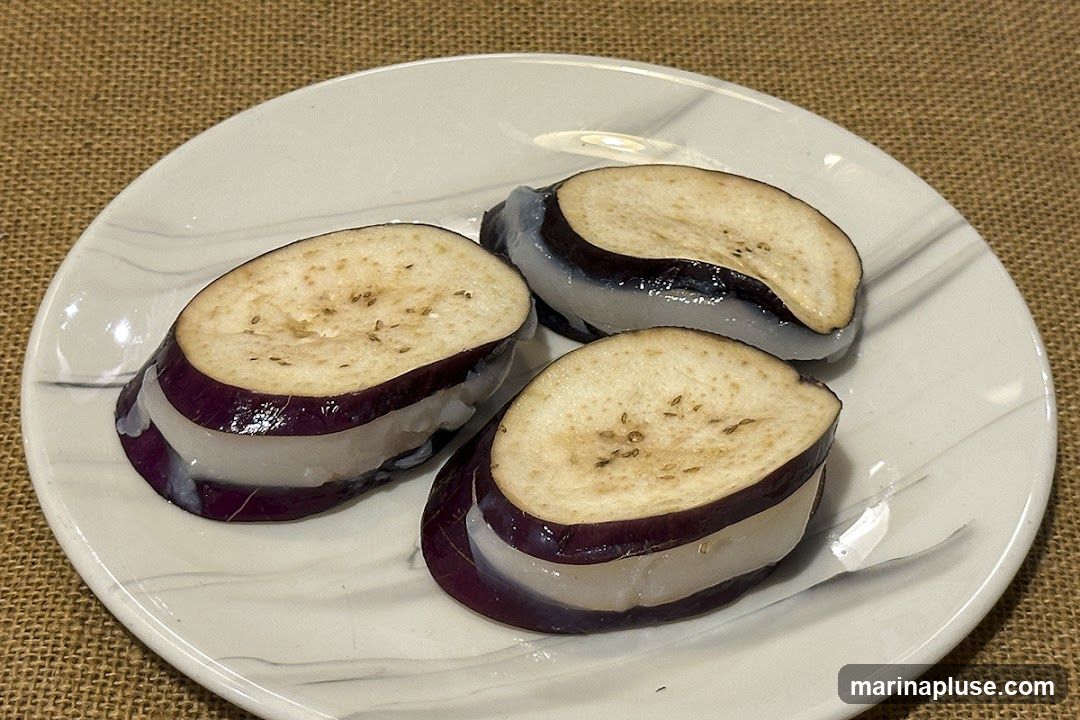 Three slices of eggplant are layered with a white filling and served on a white plate with a marbled pattern. The eggplant slices are cut crosswise, revealing the purple skin and the lighter flesh with visible seeds. The white filling appears to be soft and slightly translucent. The plate is set on a textured brown surface.