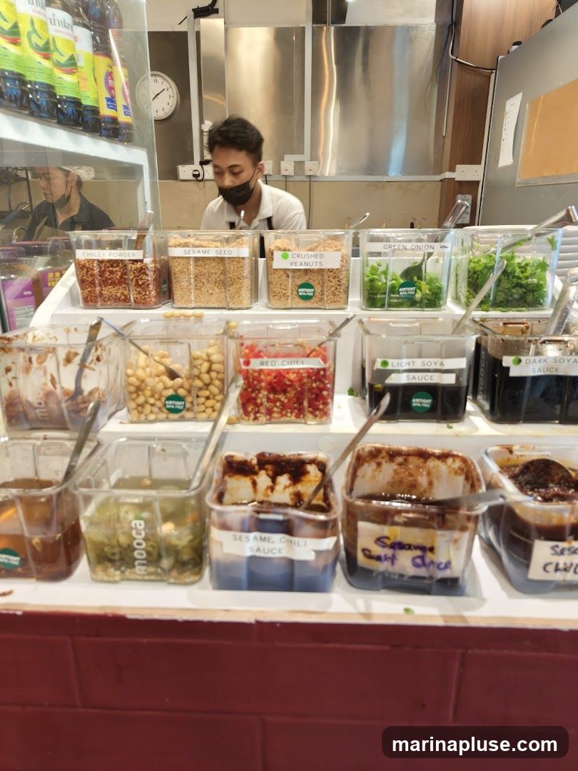 A condiment bar with various ingredients and sauces in clear containers. Ingredients include chili powder, sesame seeds, crushed peanuts, green onions, and red chilies. Sauces include light soy sauce, dark soy sauce, and sesame chili sauce. A person in the background is wearing a mask and appears to be working behind the counter.