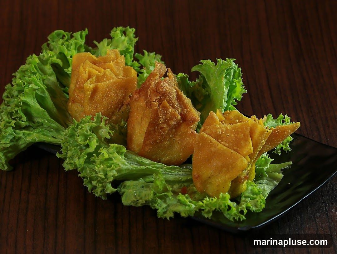 Three golden fried wontons are presented on a bed of crisp green lettuce leaves, arranged on a black square plate. The wontons are folded into a decorative, rose-like shape. The background is a dark, textured wooden surface.