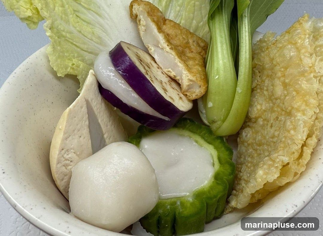 A close-up shot of a bowl filled with various ingredients, including lettuce, sliced eggplant with white filling, fried tofu, bok choy, a fish ball, stuffed bitter melon, and fried tofu skin.