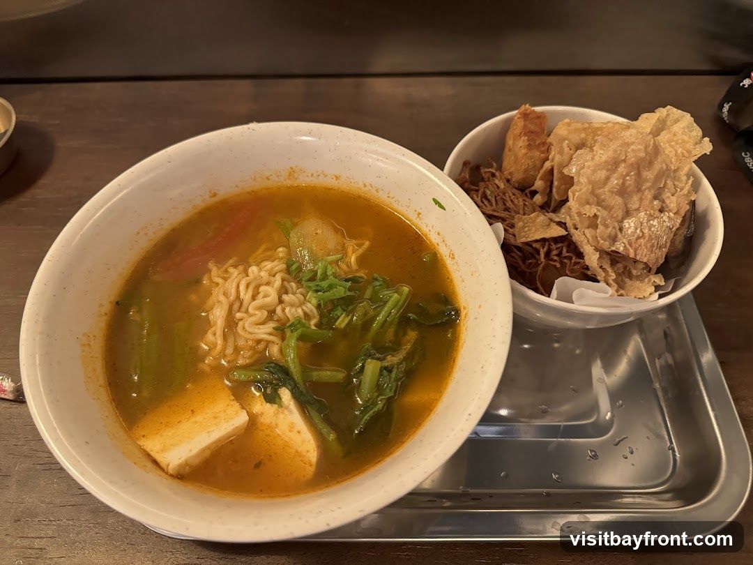 A bowl of noodle soup with tofu and vegetables is served on a metal tray. A separate bowl contains fried snacks, including crispy wonton wrappers and fried noodles.