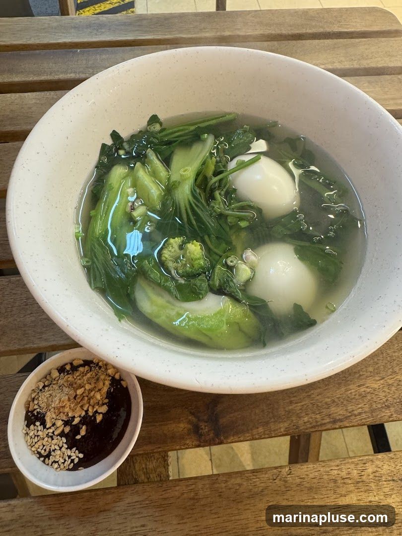 A bowl of clear soup filled with bok choy, other leafy greens, and two white dumplings. A small dish of dark sauce topped with sesame seeds and crushed nuts is placed next to the bowl. The food is served on a wooden table.