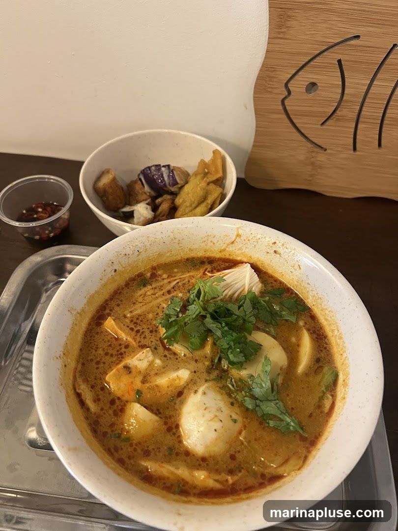 A bowl of noodle soup with various ingredients such as tofu, fish balls, and cilantro is served on a metal tray. A small bowl of fried items, including eggplant and wontons, is placed behind the soup. A small container of dipping sauce is to the left. A wooden coaster with a fish design is visible on the right.