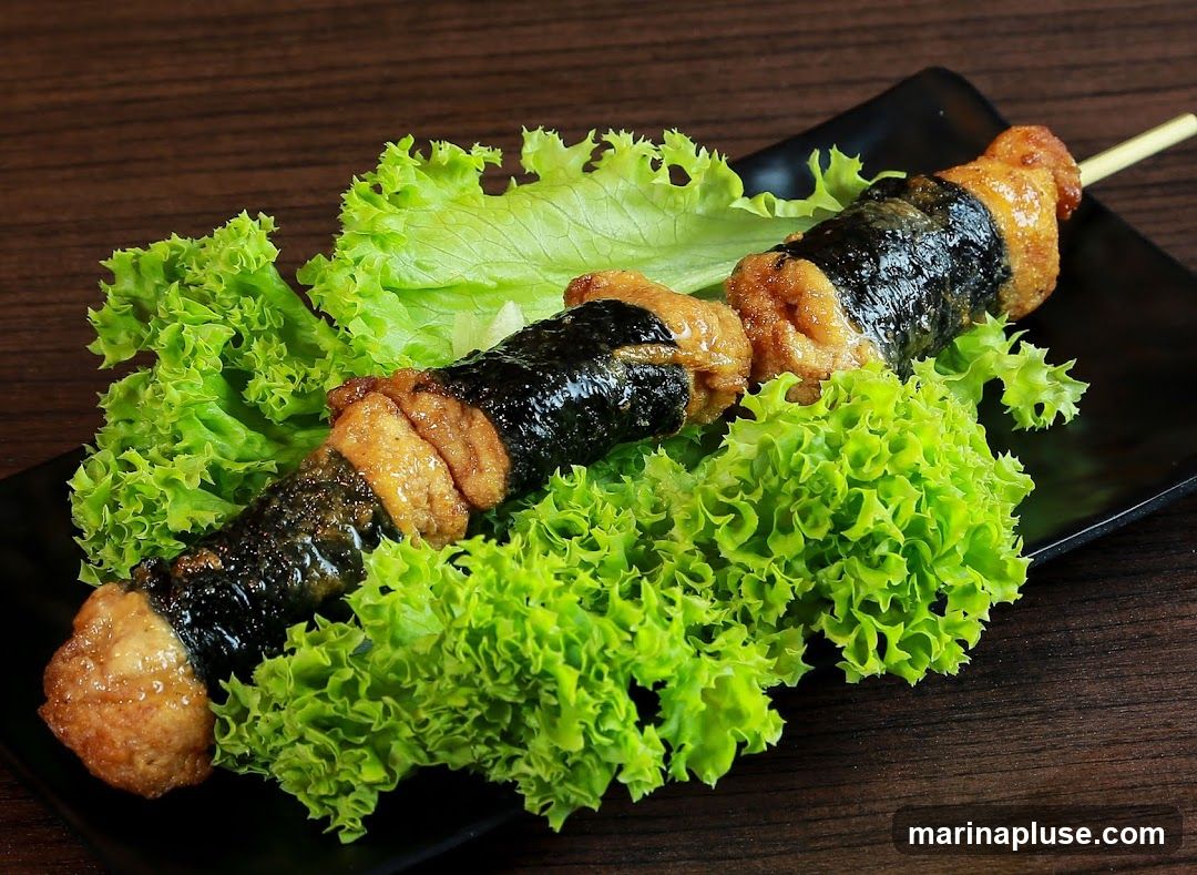 A close-up shot of a skewer with pieces of food wrapped in dark seaweed and interspersed with golden-brown fried pieces. The skewer is presented on a black plate, nestled amongst vibrant green lettuce leaves. The background is a dark, textured wooden surface.