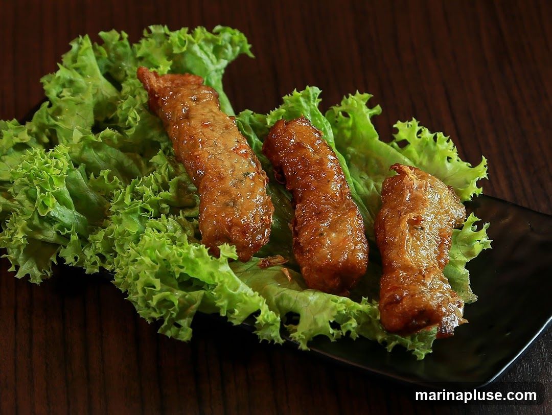 Three golden-brown fried meat rolls are arranged on a bed of fresh green lettuce leaves, served on a black rectangular plate. The rolls appear to be coated in a glossy sauce, and the lettuce provides a crisp contrast. The background is a dark, textured wooden surface.