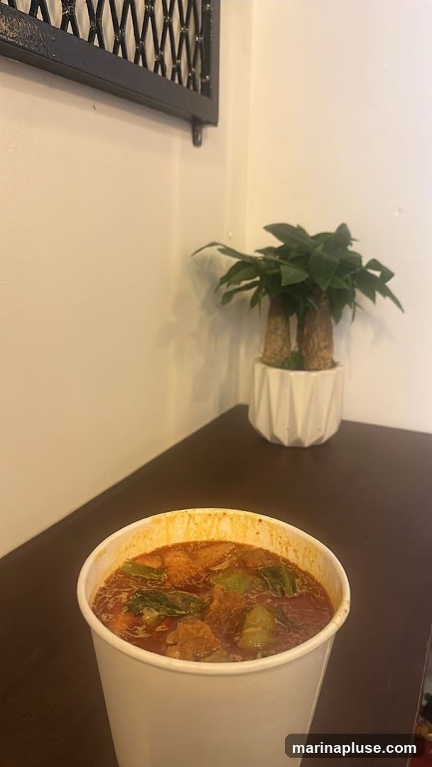 A white paper container filled with a hearty soup is placed on a dark wooden table. The soup appears to have chunks of meat and vegetables in a rich, reddish-brown broth. In the background, a white geometric planter holds a green plant, and a black decorative metal grate hangs on the wall.