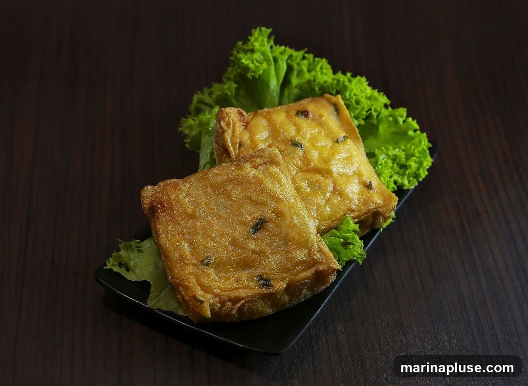 Two golden-brown, square-shaped slices of fried tofu are served on a black plate with a garnish of bright green lettuce. The tofu appears to have small green specks, possibly herbs or spices, embedded within its crispy exterior. The dark, wooden background provides a contrasting backdrop, highlighting the food.