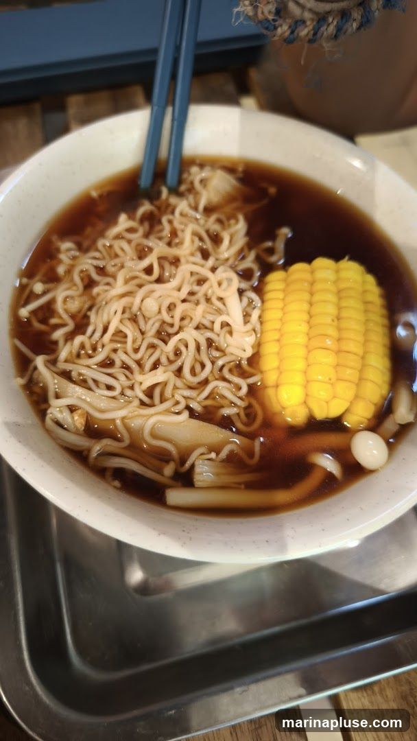 A close-up shot of a bowl of noodle soup. The soup contains instant noodles, enoki mushrooms, a segment of corn on the cob, and what appears to be a quail egg. Blue chopsticks are resting in the bowl. The bowl is placed on a metal tray, which is on a wooden surface.