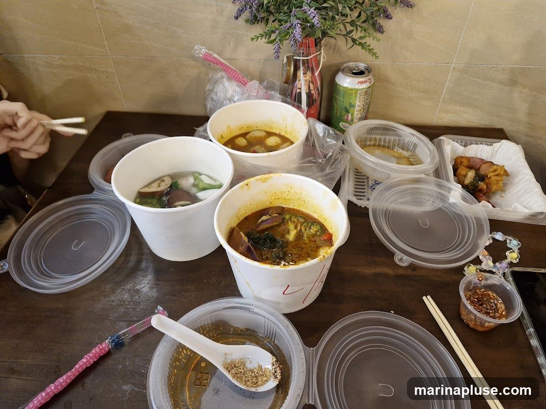 A table laden with several takeout containers of food, including bowls of soup with various ingredients like vegetables and meatballs, a container with fried food, and a small dish of sauce. Chopsticks and a spoon are visible, along with a can of drink and a decorative item. The scene suggests a casual meal enjoyed at home or a similar setting.