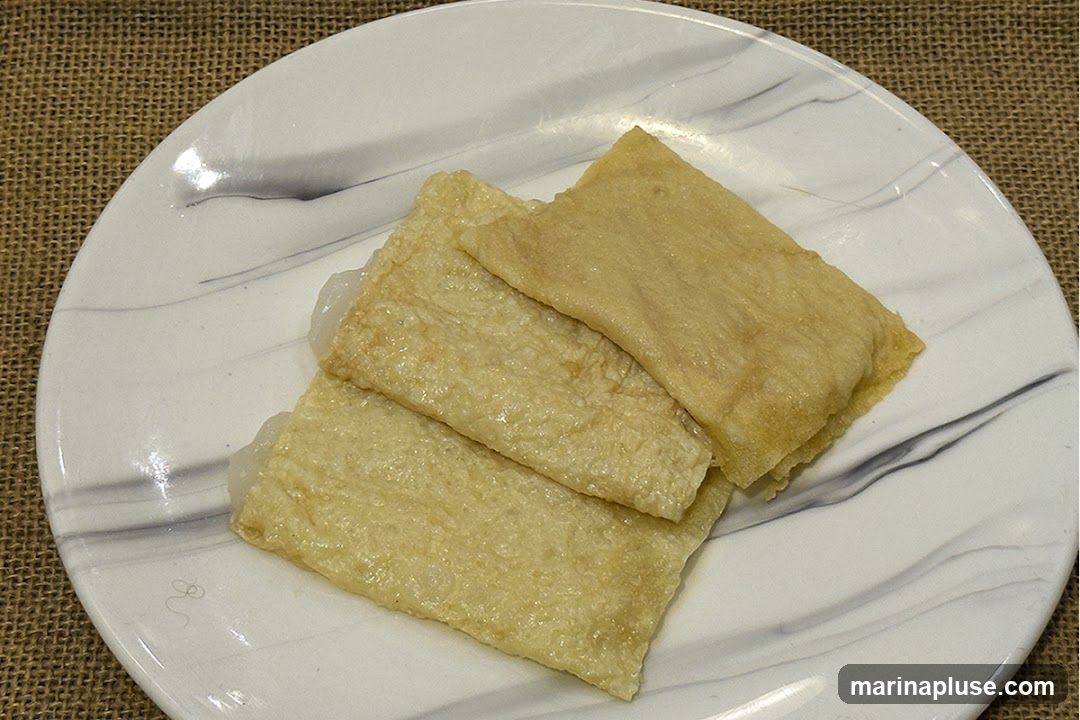 Three rectangular pieces of fried tofu skin are stacked on a white plate with a marble-like pattern. The tofu skin is golden brown and appears slightly crispy. A white filling is visible peeking out from the edges of the wraps. The plate is set on a textured brown surface, possibly burlap.