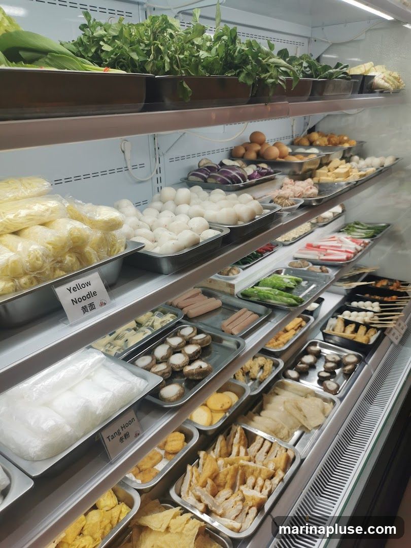 A refrigerator display case filled with various ingredients, likely for hot pot or a similar dish. The top shelf holds fresh green vegetables. Below, trays contain items such as yellow noodles, white fish balls, sliced eggplant, eggs, sausages, mushrooms, tofu, and various fried items. Labels indicate 'Yellow Noodle' and 'Tang Hoon'.