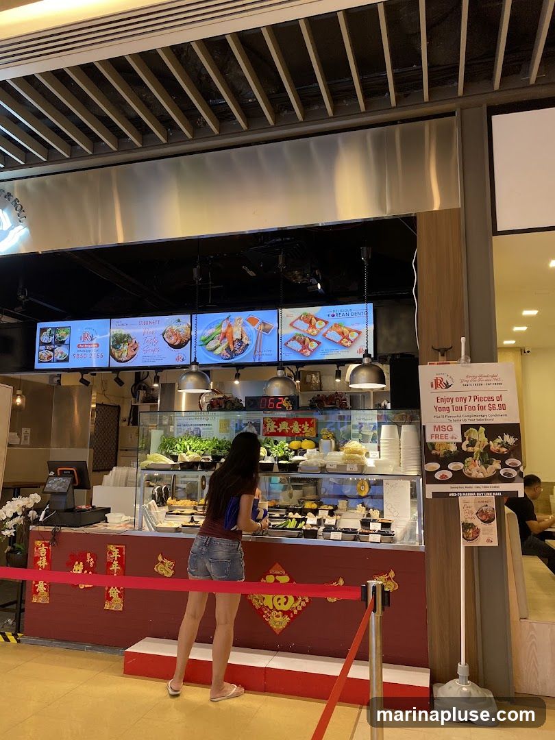 A food stall counter with a display of various food items and a digital menu board above. A woman is standing at the counter, looking at the food. There is a promotional poster on the right advertising Yong Tau Foo.