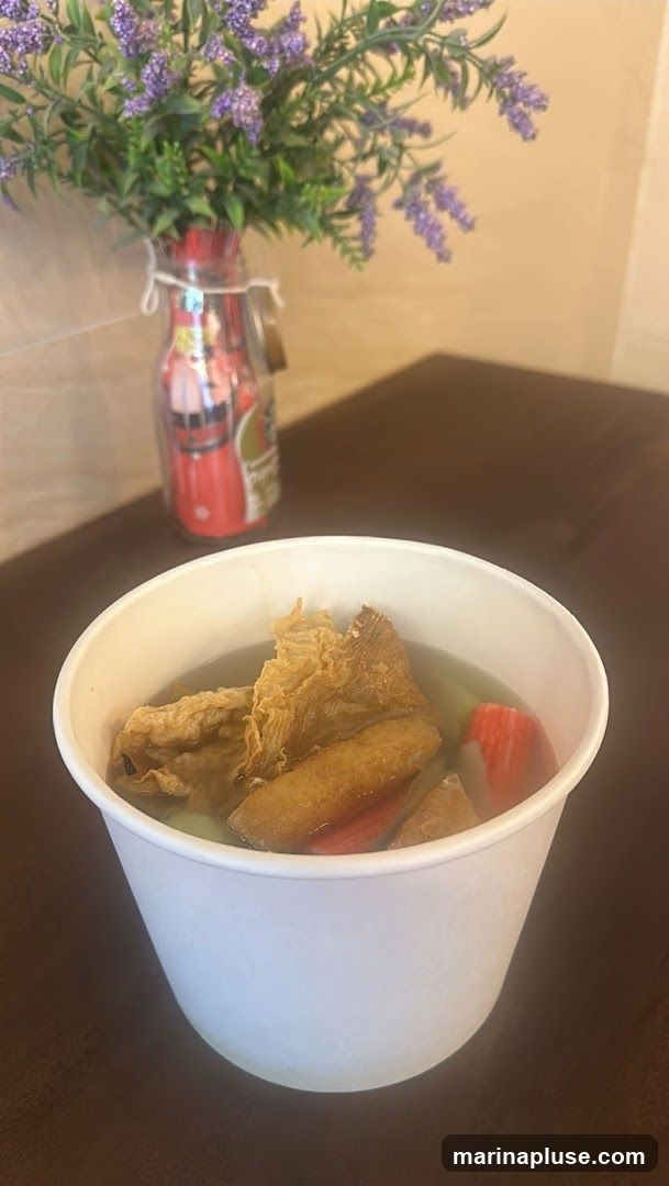 A white paper bowl filled with soup and various ingredients, including fried tofu, fish cakes, and what appears to be imitation crab sticks. The bowl is placed on a dark brown surface. In the background, a vase filled with artificial purple flowers adds a decorative touch. The vase itself is red and decorated with images.