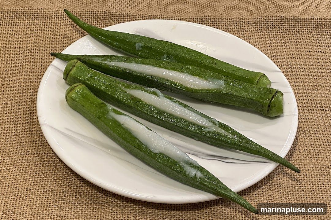 Four pieces of okra are arranged on a white plate with a marble pattern. Each piece of okra has been cut lengthwise and filled with a white, creamy substance. The plate is set on a textured brown surface, possibly burlap.