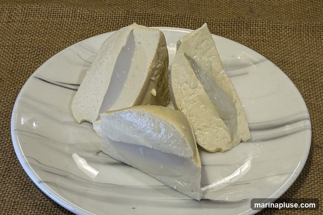 Three slices of tofu are arranged on a white plate with a marbled gray pattern. The tofu appears to be firm and has a slightly creamy interior. The plate is set on a textured brown surface, possibly burlap.