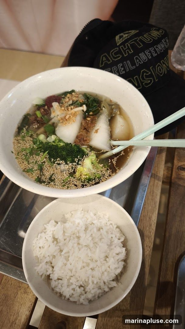 A bowl of noodle soup with vegetables and a side of white rice. A black baseball cap with text is visible in the background.