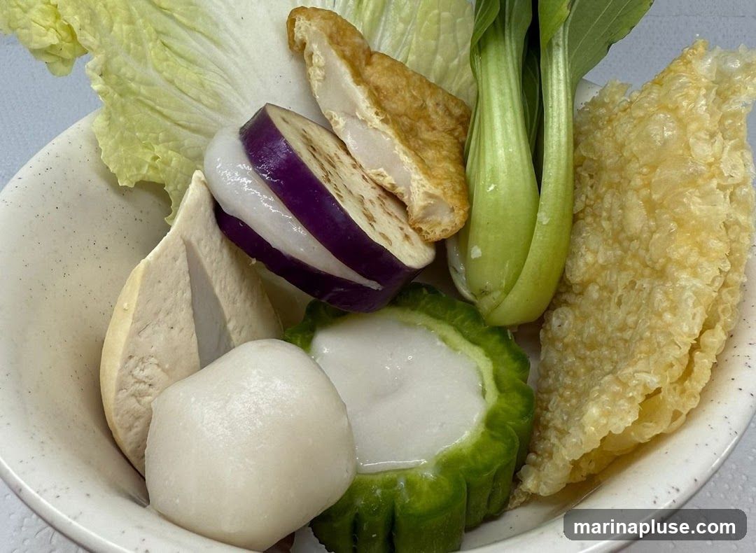 A close-up shot of a bowl filled with various ingredients, likely for a hot pot or soup. Visible items include napa cabbage, sliced eggplant with white filling, fried tofu, bok choy, a fish ball, and a slice of bitter melon stuffed with white paste. A piece of crispy fried bean curd skin is also present on the right side.