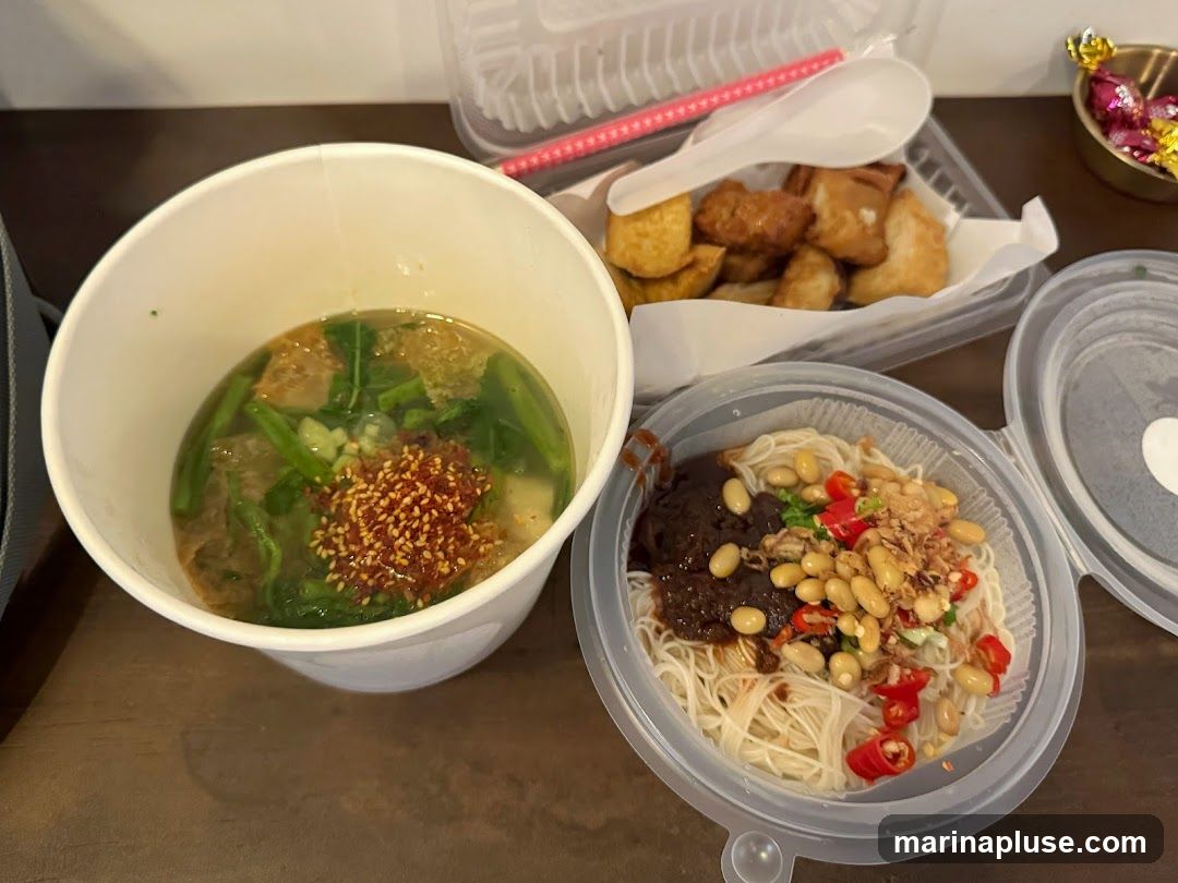 A close-up view of a takeaway meal, featuring a bowl of noodles with various toppings including soybeans, chili slices, and fried shallots, alongside a cup of soup with greens and chili flakes. A separate container holds several pieces of fried food, possibly tofu or fish cakes. A pink straw and a white spoon are visible in the background, along with a small dish of wrapped candies.