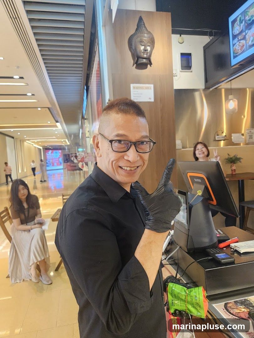A man in a black shirt and black gloves is smiling and giving a thumbs up while standing behind a counter. He is wearing glasses. In the background, a woman is smiling and making a peace sign. There is a Buddha head decoration on the wall behind them. A digital menu board is visible above the counter.