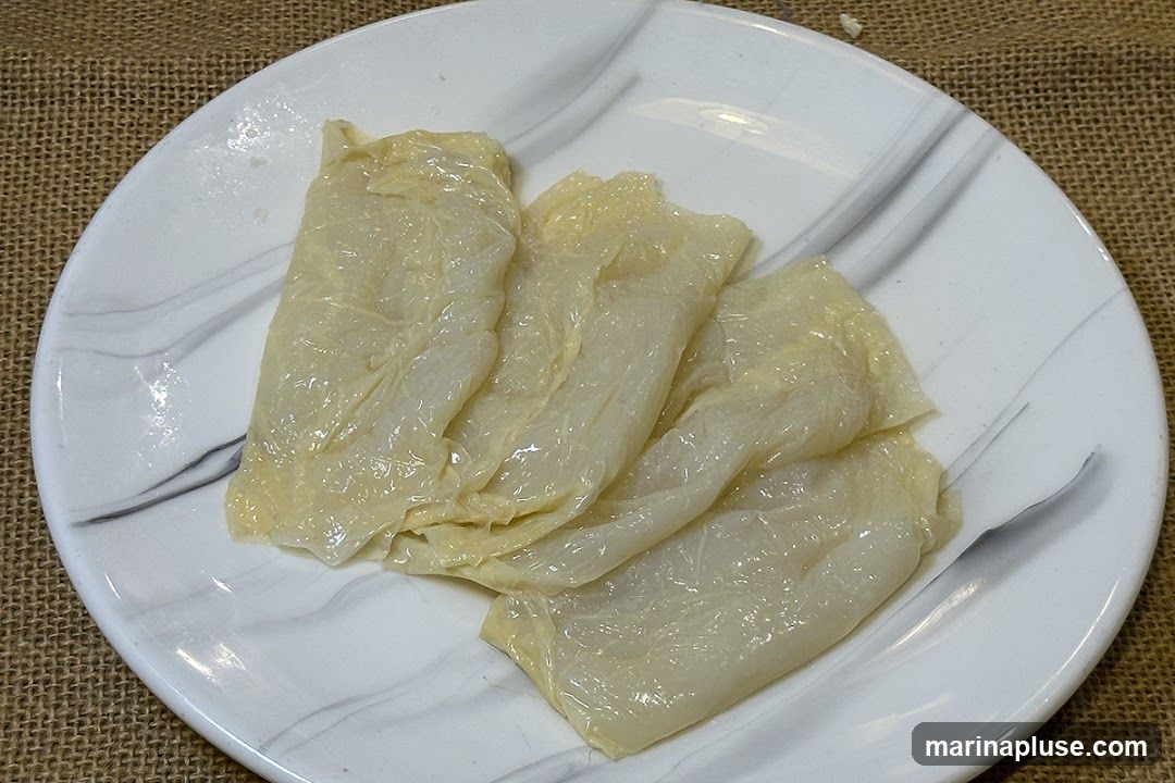 A close-up shot of several pieces of tofu skin, also known as yuba or bean curd skin, arranged on a white plate with a marble-like pattern. The tofu skin is pale yellow and appears slightly translucent and wrinkled. The plate is set on a textured brown surface, possibly burlap.