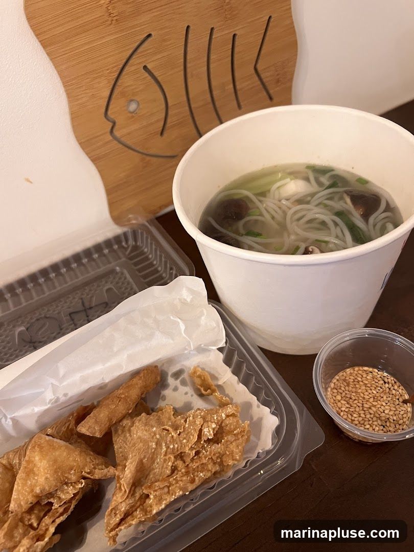 A close-up view of a takeout meal, featuring a bowl of noodle soup with mushrooms and greens, a container of fried items, and a small dish of sesame seeds. A wooden cutting board with a fish design is visible in the background.