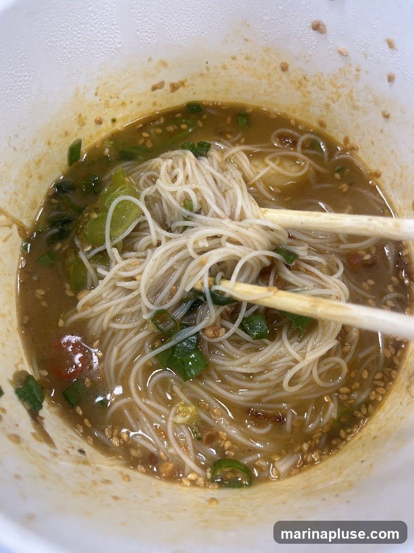 A close-up shot of a bowl of noodle soup. Thin white noodles are submerged in a savory broth, garnished with chopped green onions, sesame seeds, and what appear to be pieces of chili and garlic. Chopsticks are holding a portion of the noodles. The bowl has a white, textured rim, and some broth has splattered on the inside.