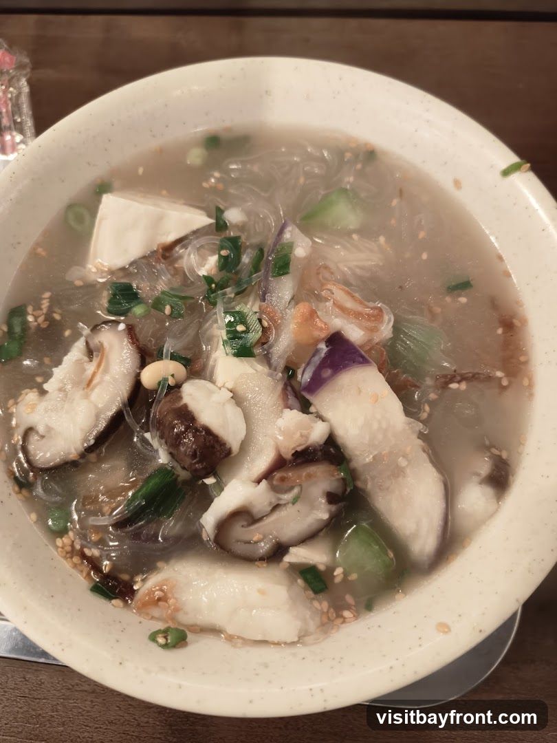 A close-up shot of a bowl of soup filled with thin noodles, cubes of tofu, sliced shiitake mushrooms, pieces of eggplant, and chopped green onions. Sesame seeds are sprinkled throughout the broth.