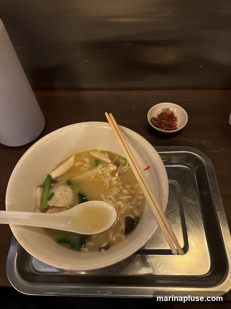 A bowl of noodle soup with various ingredients including mushrooms, greens, and what appear to be fish balls, served with chopsticks and a spoon. A small dish of chili paste is on the side. The meal is presented on a metal tray.