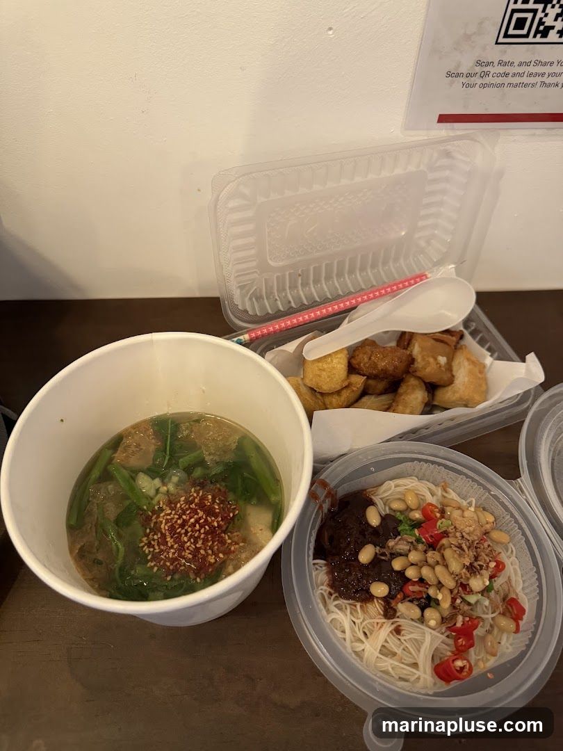 A close-up shot of takeout food containers on a wooden surface. In the foreground, a white paper bowl contains soup with greens and a sprinkle of red chili flakes. To the right, a clear plastic container holds thin noodles topped with a dark sauce, soybeans, chili slices, and fried shallots. Behind these, another clear plastic container holds fried food items. A QR code and text encouraging customers to scan, rate, and share their opinions are visible on the wall in the background.