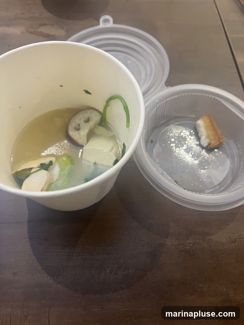 A close-up overhead view of two white takeout containers on a dark wooden surface. The larger container, a paper cup, is mostly full of a light-colored broth with various ingredients including slices of eggplant, tofu, and greens. The smaller, clear plastic container has a lid slightly ajar and contains a single piece of fried food.