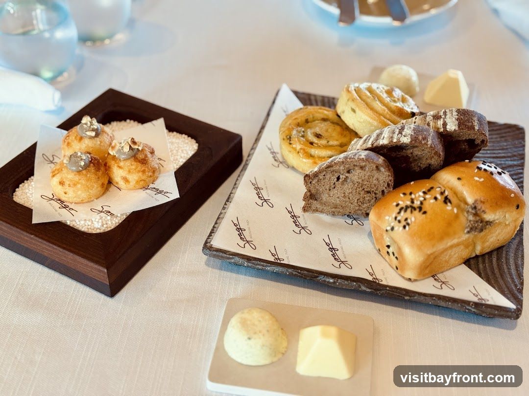A bread basket with various types of bread, rolls, and butter. There are also three small savory puffs topped with a grey spread and nuts on a separate wooden tray. The bread is served on parchment paper with a logo.