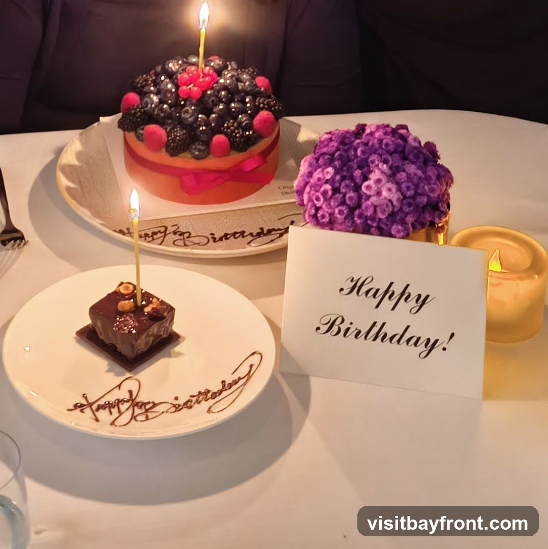 A table setting with two birthday desserts, a larger cake topped with berries and a smaller chocolate pastry. Both have lit candles. A "Happy Birthday!" card, a small bouquet of purple flowers, and a lit candle are also on the table.