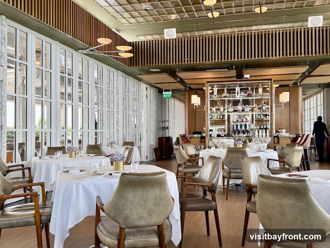 The interior of a restaurant with tables set for dining. The room features large windows, a bar area with shelves stocked with bottles, and comfortable seating. A person in a suit is visible near the bar.
