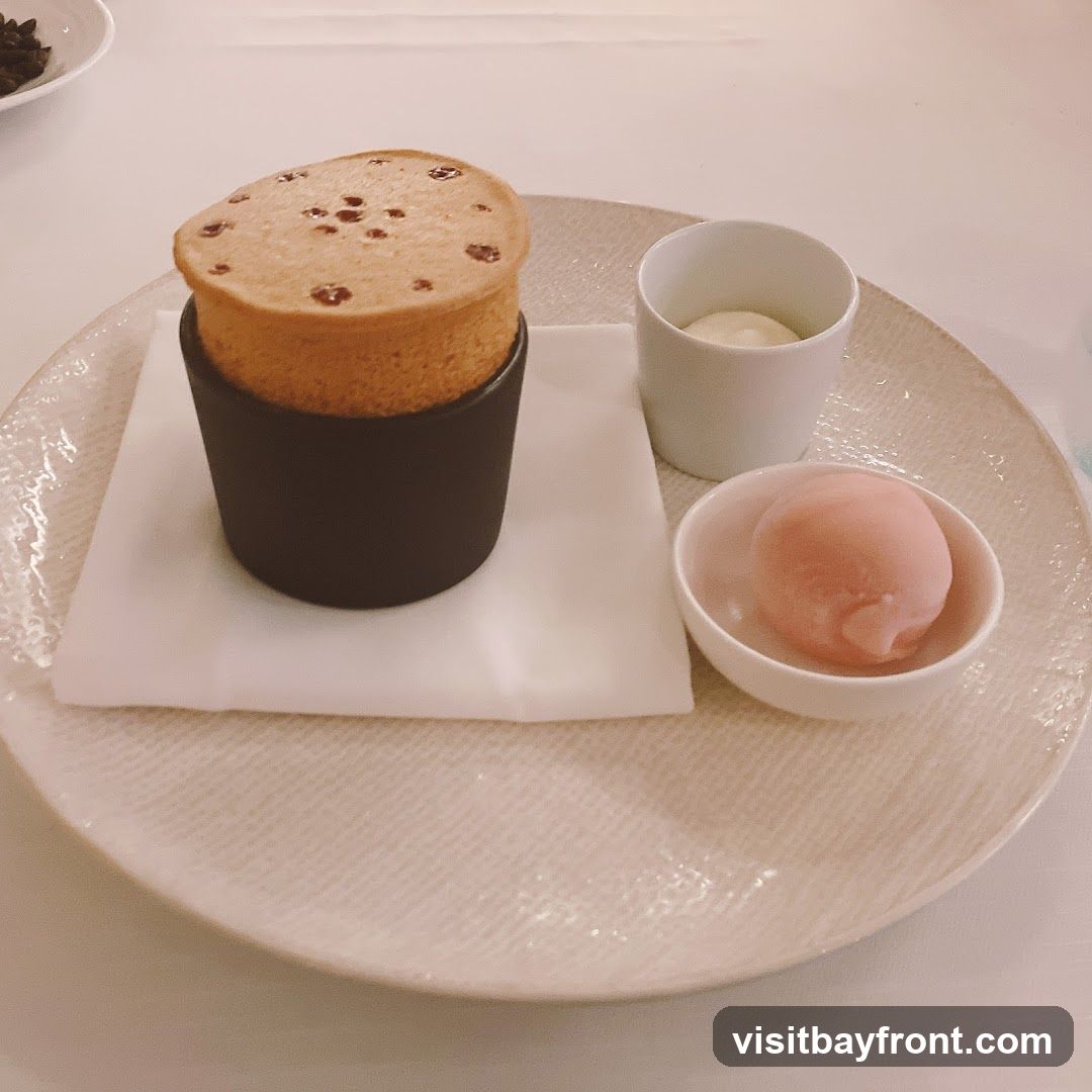 A close-up shot of a dessert on a white plate. The dessert features a soufflé in a dark ramekin, a scoop of pink sorbet in a small white bowl, and a dollop of cream in a white cup. The soufflé has a golden-brown top with small red dots.
