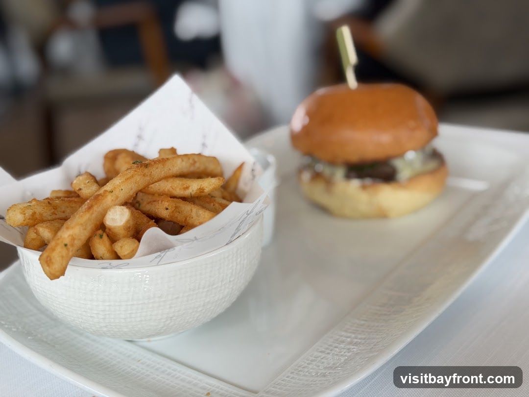 A close-up shot of a slider burger and a bowl of french fries on a white plate. The burger has a golden bun and is topped with melted cheese. The fries are golden brown and seasoned. The background is blurred, showing hints of a restaurant interior.
